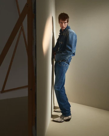 Full-length portrait of a male model wearing a denim jacket and jeans, standing against a neutral studio wall by Giuseppe Triscari.
