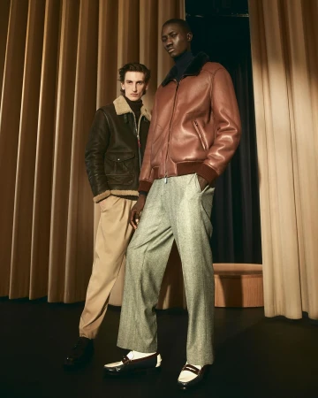 Two male fashion models pose in studio wearing shearling-collared leather jackets, wide trousers, and loafers against a curtain backdrop.