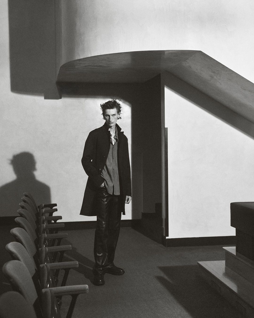 A man in a dark coat and pants poses in a modern building with rows of chairs and geometric architectural elements in a fashion shot.