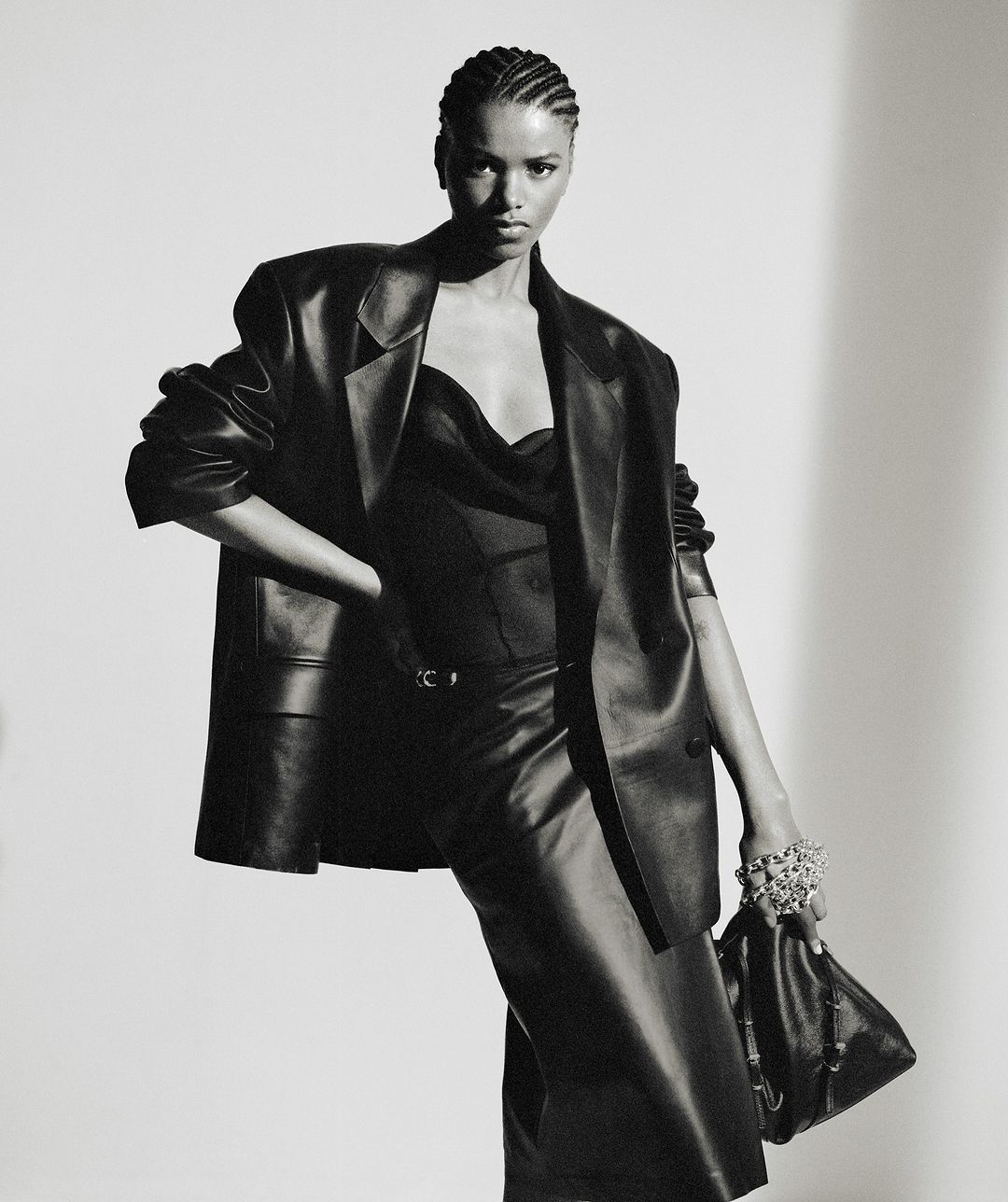 A model in a black leather outfit poses in a studio, wearing an oversized blazer, top, and skirt, accessorized with a bracelet and handbag.
