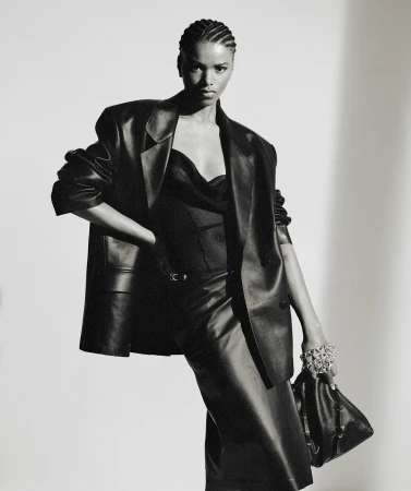 A model in a black leather outfit poses in a studio, wearing an oversized blazer, top, and skirt, accessorized with a bracelet and handbag.