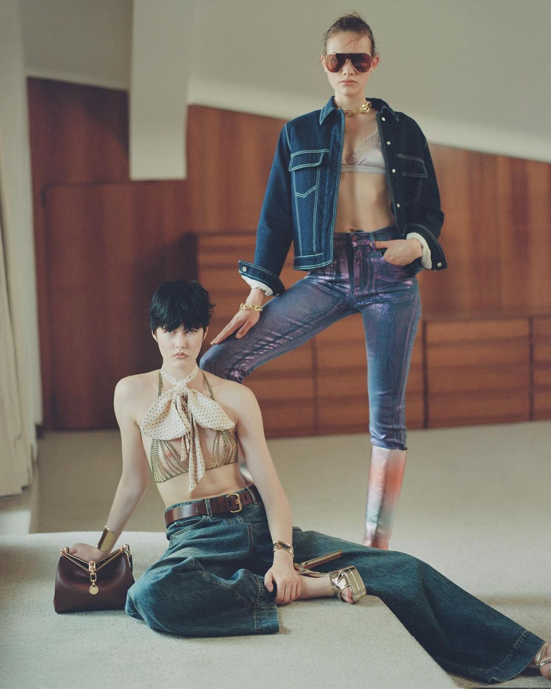 Two women in retro fashion pose indoors. One sits in jeans and halter top with a handbag, while the other stands in denim and metallic pants.