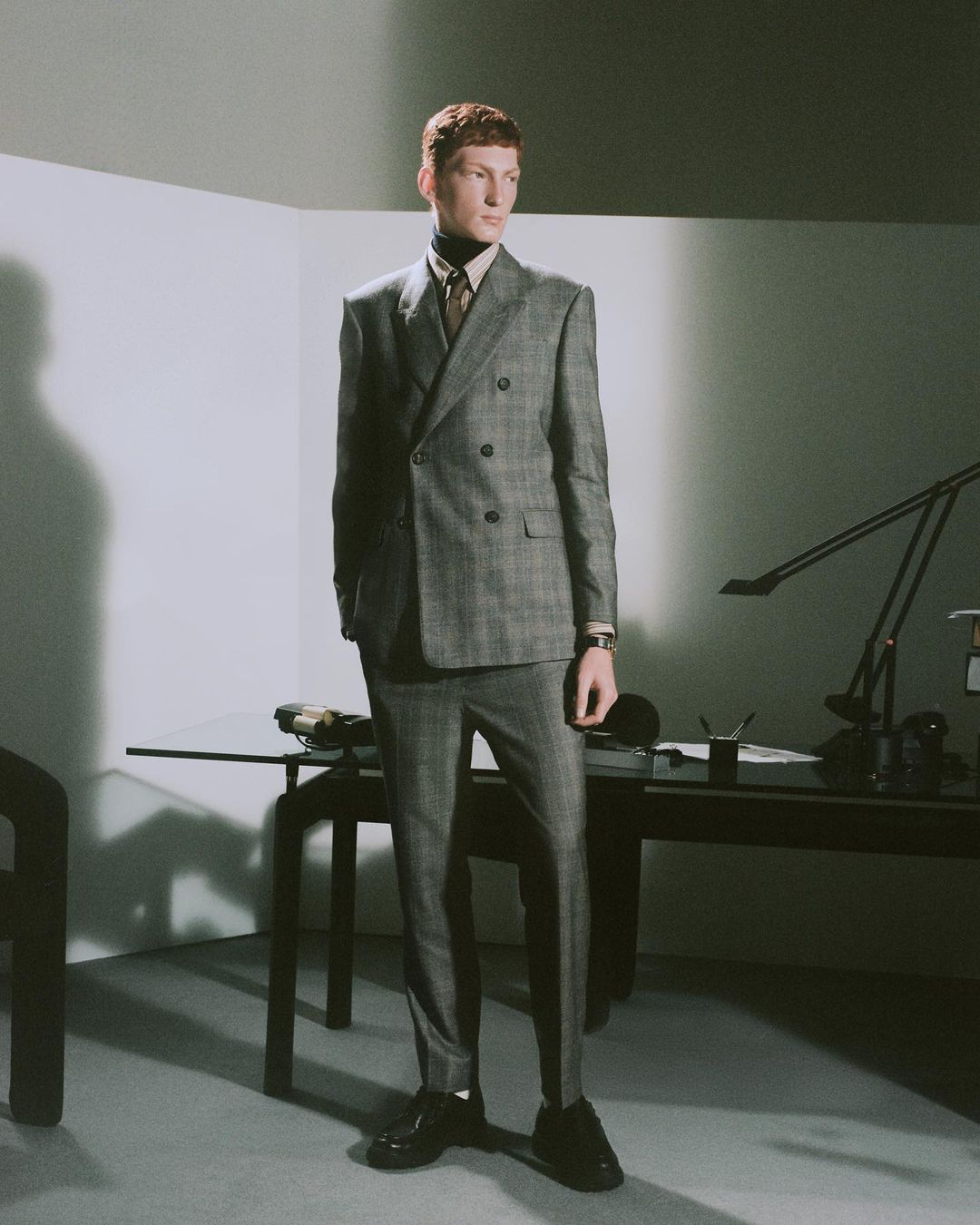 Man in a grey plaid double-breasted suit poses indoors by a desk with a lamp in a modern, naturally lit interior space.