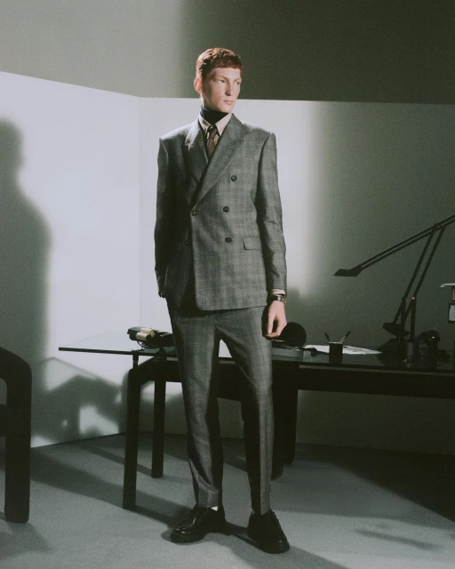 Man in a grey plaid double-breasted suit poses indoors by a desk with a lamp in a modern, naturally lit interior space.