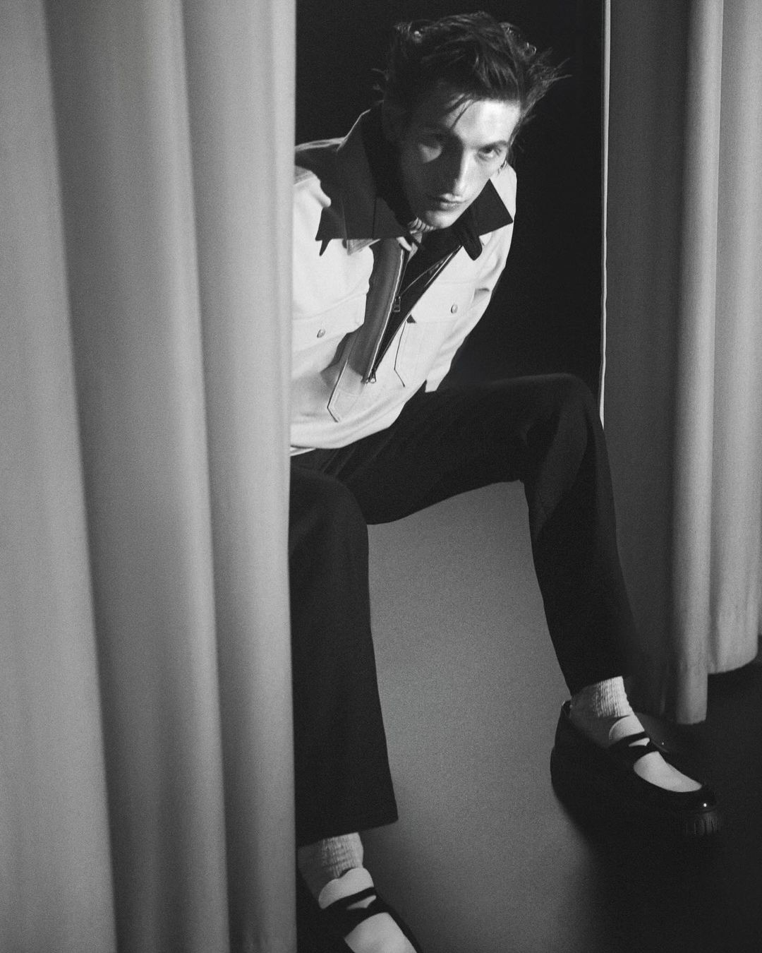 Monochrome portrait of a man sitting between draped curtains wearing a light jacket, dark trousers, and buckled shoes
