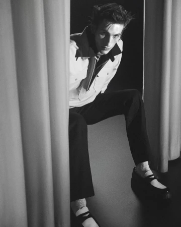 Monochrome portrait of a man sitting between draped curtains wearing a light jacket, dark trousers, and buckled shoes