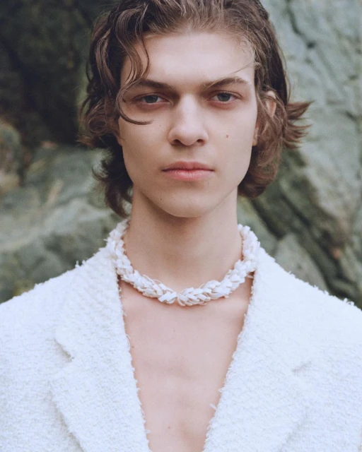 Close-up studio portrait of a young adult male with wavy brown hair wearing a white blazer and a white braided necklace, outdoors.