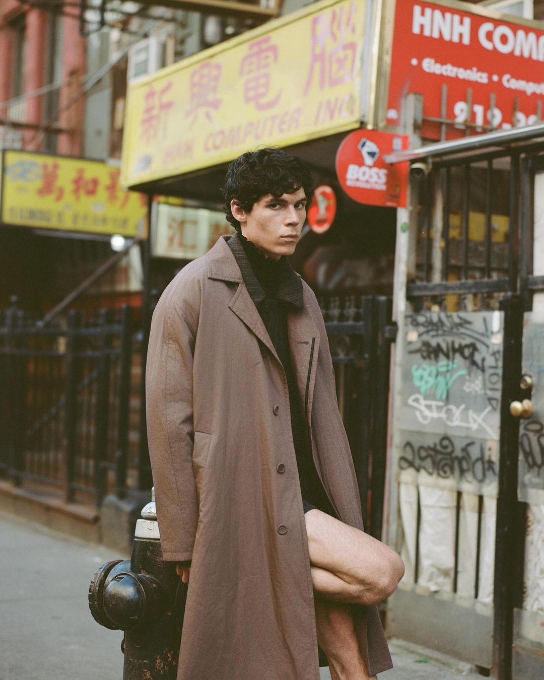 A brunette male model poses in a brown coat in an urban setting with signage in the background, photographed in a medium shot.