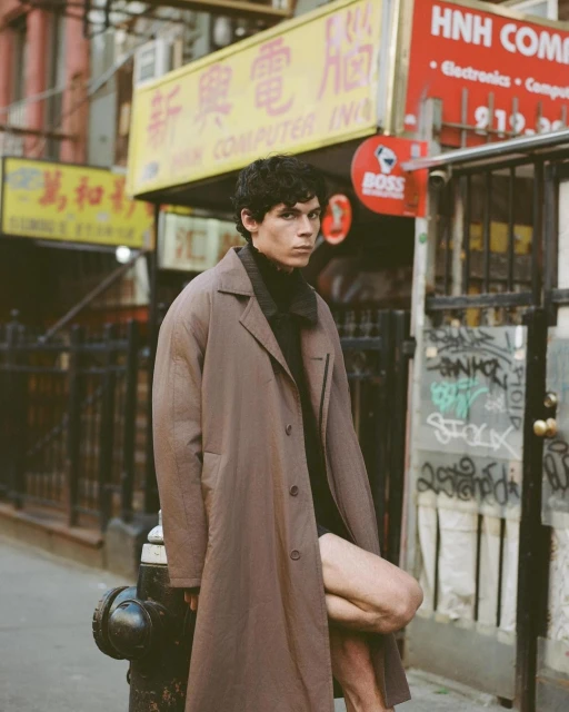 A brunette male model poses in a brown coat in an urban setting with signage in the background, photographed in a medium shot.