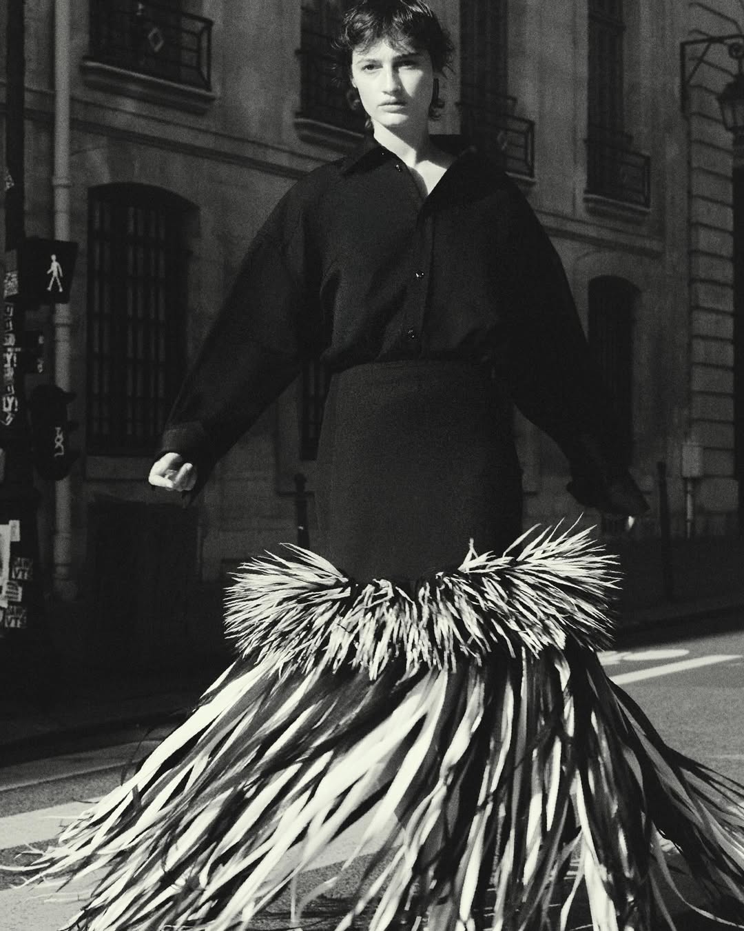 Monochrome fashion shot of an adult woman in dark shirt and statement skirt made of fronds on a city street with stone buildings.