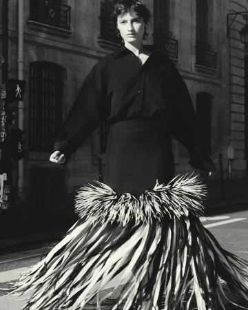 Monochrome fashion shot of an adult woman in dark shirt and statement skirt made of fronds on a city street with stone buildings.