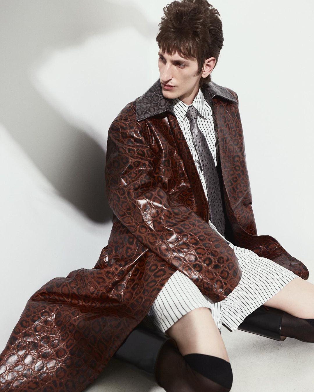 Man in a brown faux crocodile leather coat, striped shirt, black boots and socks poses against a gradient white background.