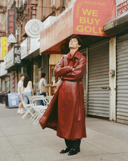 A stylish adult male stands confidently on a city sidewalk wearing a red leather trench coat, a matching shirt, and black trousers.