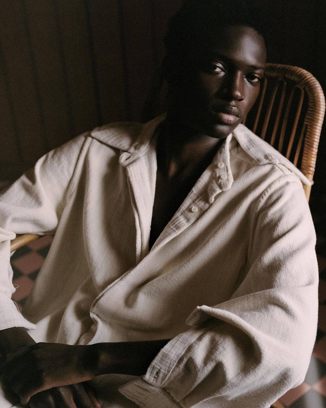 Portrait of a dark-skinned adult male indoors, wearing an off-white button-down shirt, seated in a wicker chair with patterned flooring.