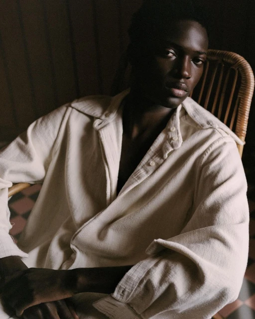 Portrait of a dark-skinned adult male indoors, wearing an off-white button-down shirt, seated in a wicker chair with patterned flooring.