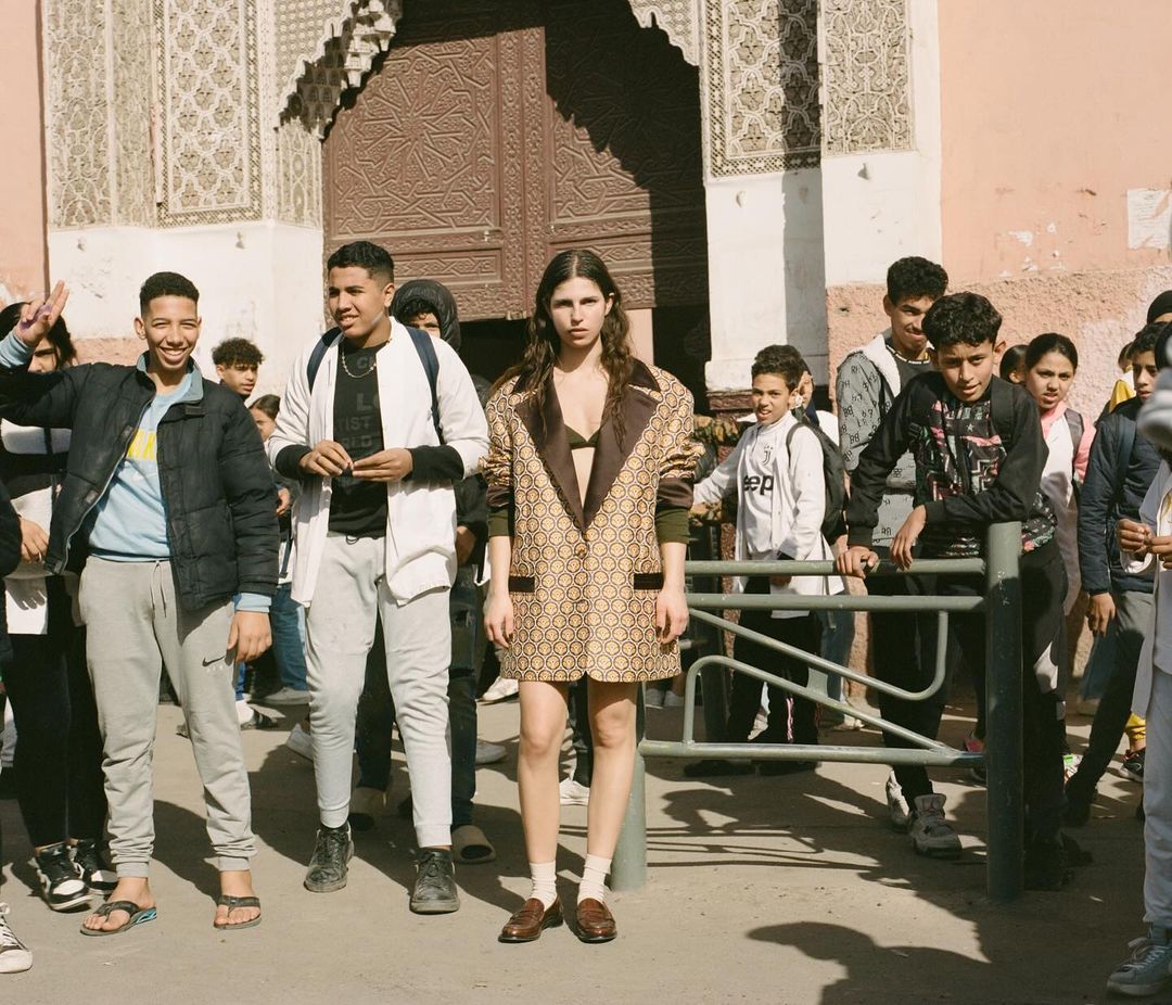 Fashion shot of a young woman in a patterned trench coat standing in front of a building with an ornate door, surrounded by people.