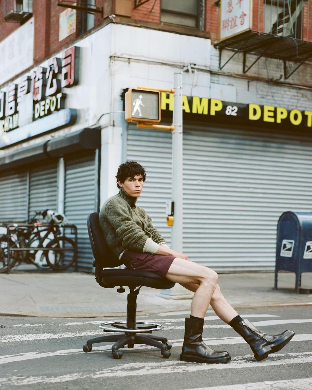 An adult male sits on an office chair in the middle of a crosswalk in a city, wearing a sweater, shorts, and boots with buildings as a backdrop.