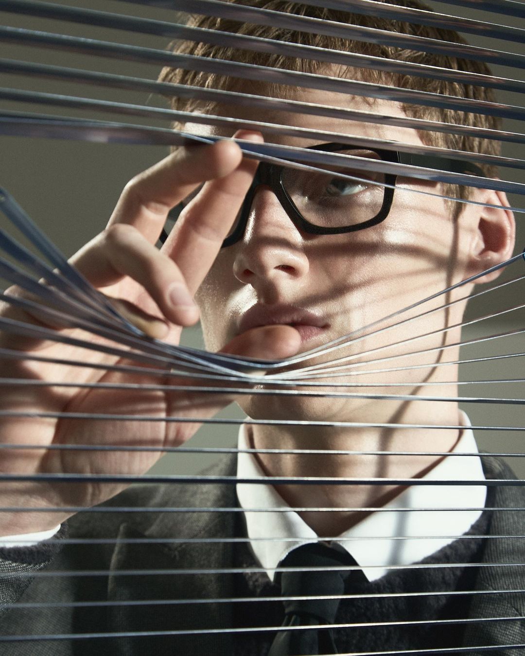 Man with glasses peers through window blinds, wearing a suit and tie in an indoor setting, with selective focus on his face.