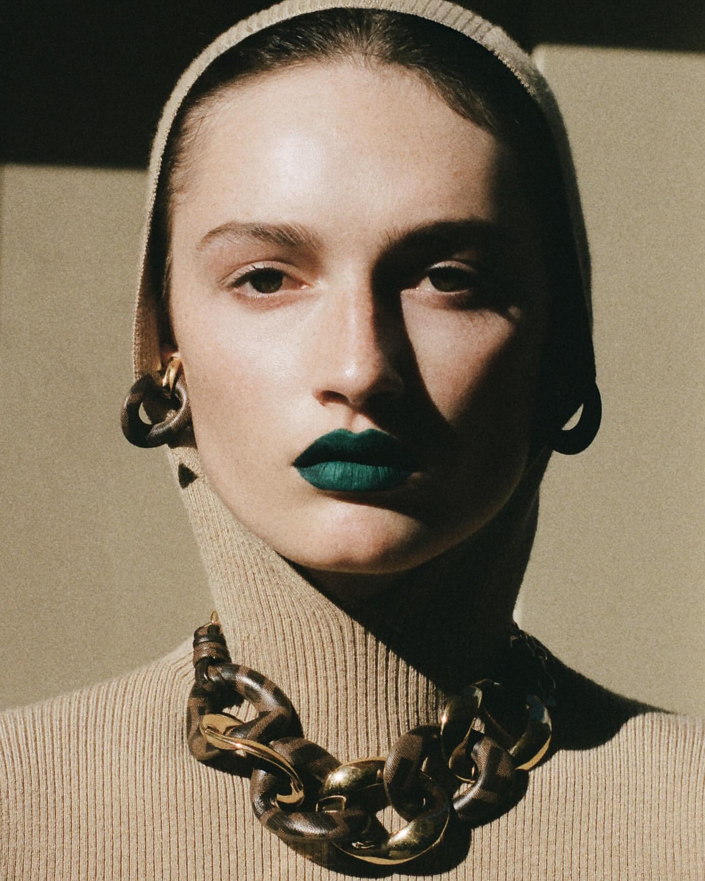 Portrait of a woman with green lipstick wearing a tan turtleneck, head covering, and brown and gold jewelry in a studio setting.