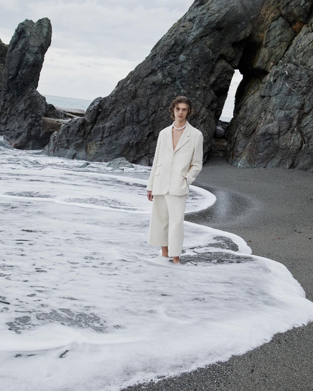 A man in an off-white suit standing in shallow seawater near dramatic rocky cliffs shot by Giuseppe Triscari