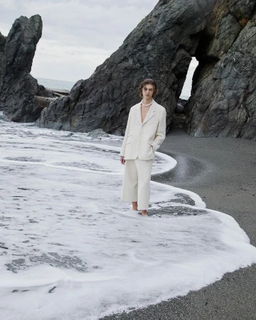 A man in an off-white suit standing in shallow seawater near dramatic rocky cliffs shot by Giuseppe Triscari