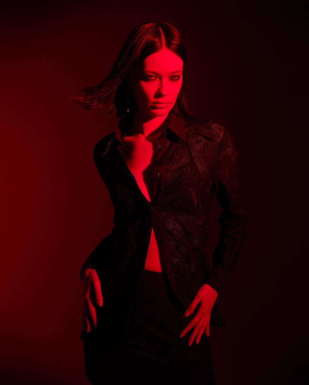 A woman with long dark hair and wearing a black shirt poses indoors in a medium shot with strong red lighting, creating a dramatic effect.