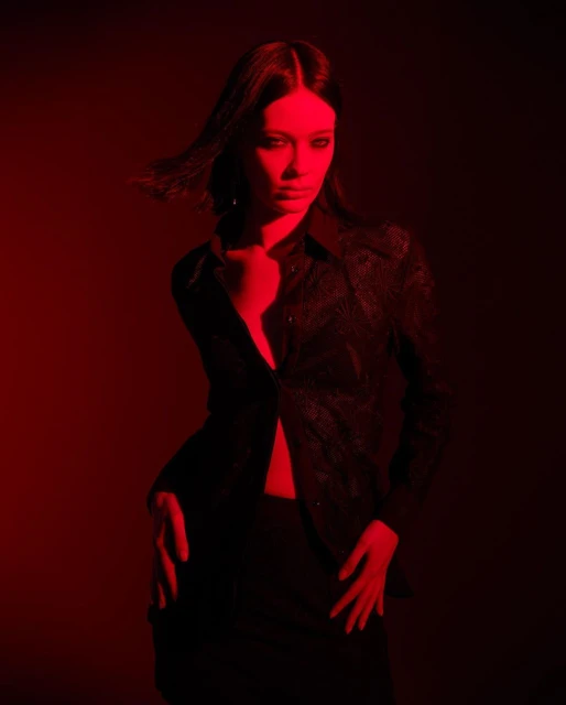 A woman with long dark hair and wearing a black shirt poses indoors in a medium shot with strong red lighting, creating a dramatic effect.