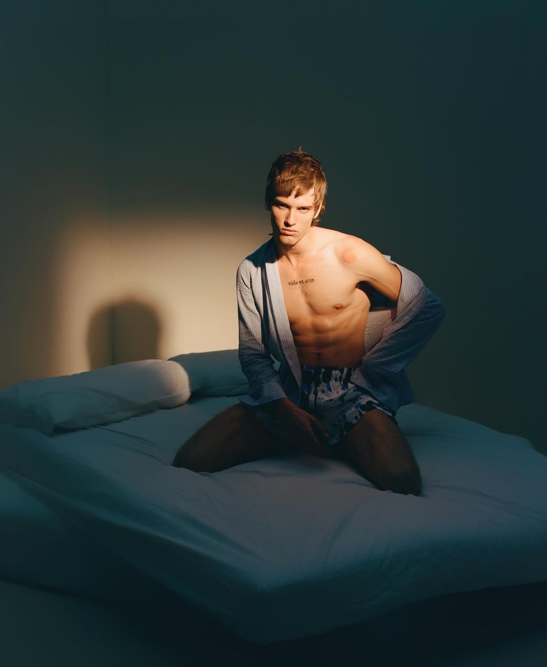 Man in grey robe and patterned shorts kneels on a white bed in soft light, looking at the camera with a neutral expression.