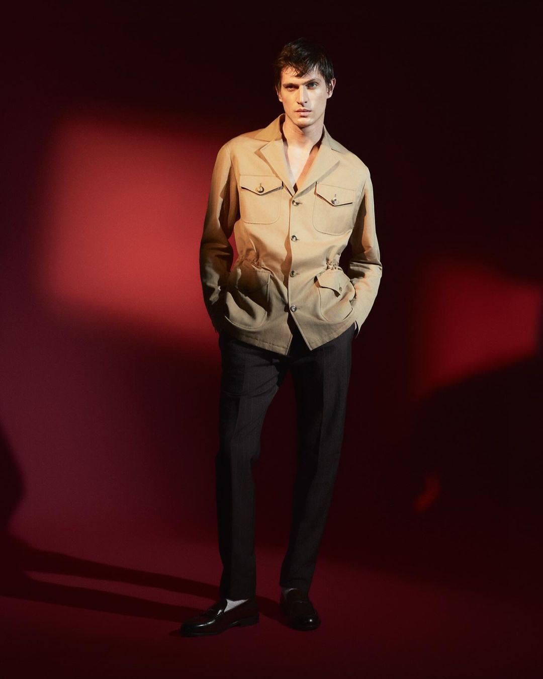 Full-length studio shot of a young adult male model posing in a beige safari jacket, dark pants, and black dress shoes against a red background.