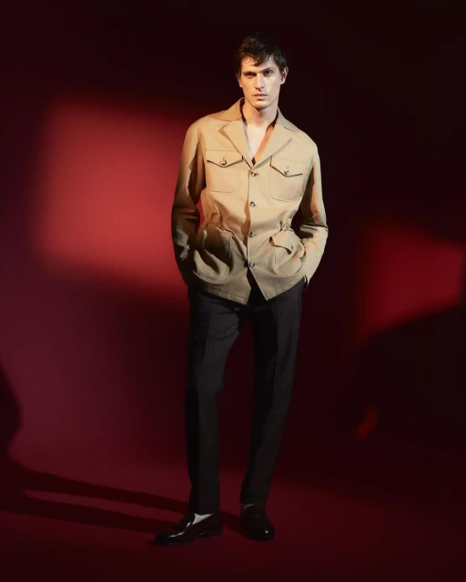 Full-length studio shot of a young adult male model posing in a beige safari jacket, dark pants, and black dress shoes against a red background.