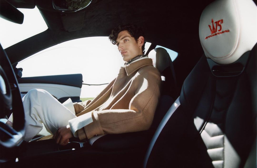A man in a tan coat sits inside a Maserati, catching sunlight, wearing white pants in the car's black and white interior.