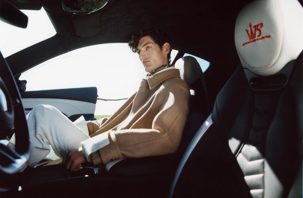 A man in a tan coat sits inside a Maserati, catching sunlight, wearing white pants in the car's black and white interior.