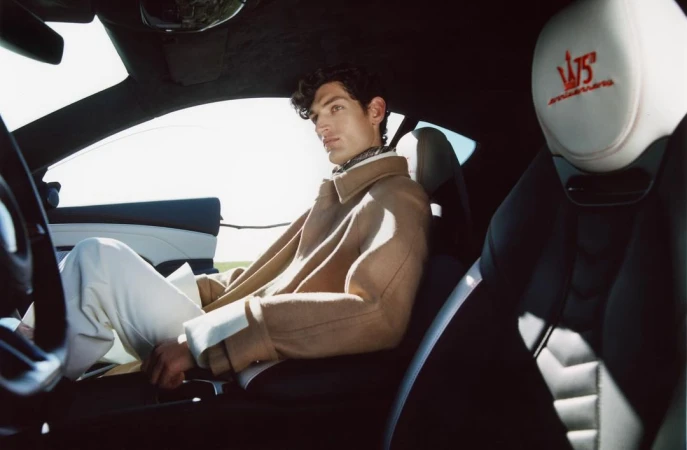 A man in a tan coat sits inside a Maserati, catching sunlight, wearing white pants in the car's black and white interior.
