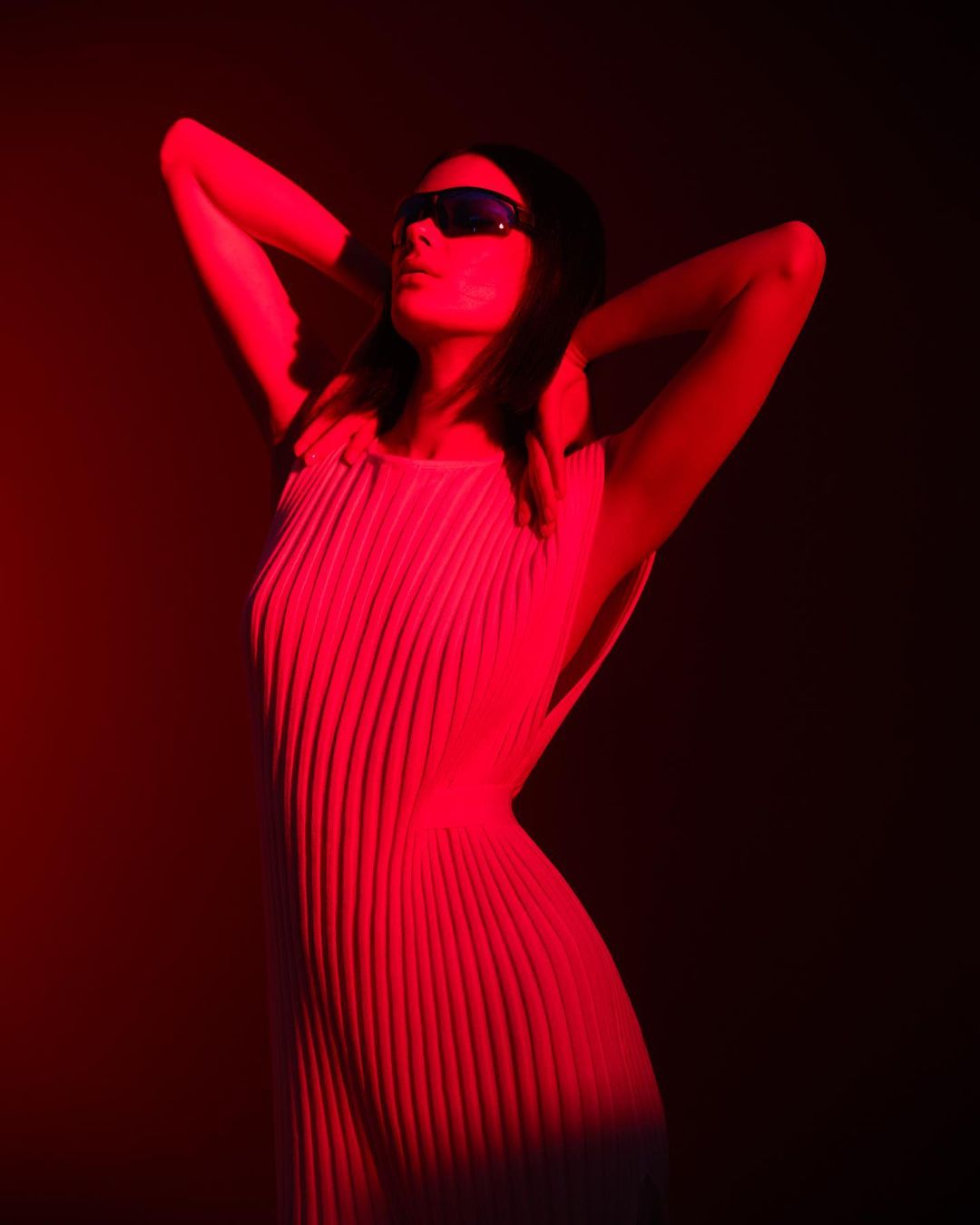 A woman in sunglasses with hands behind her head poses in front of a dark background illuminated by red light, wearing a textured dress.