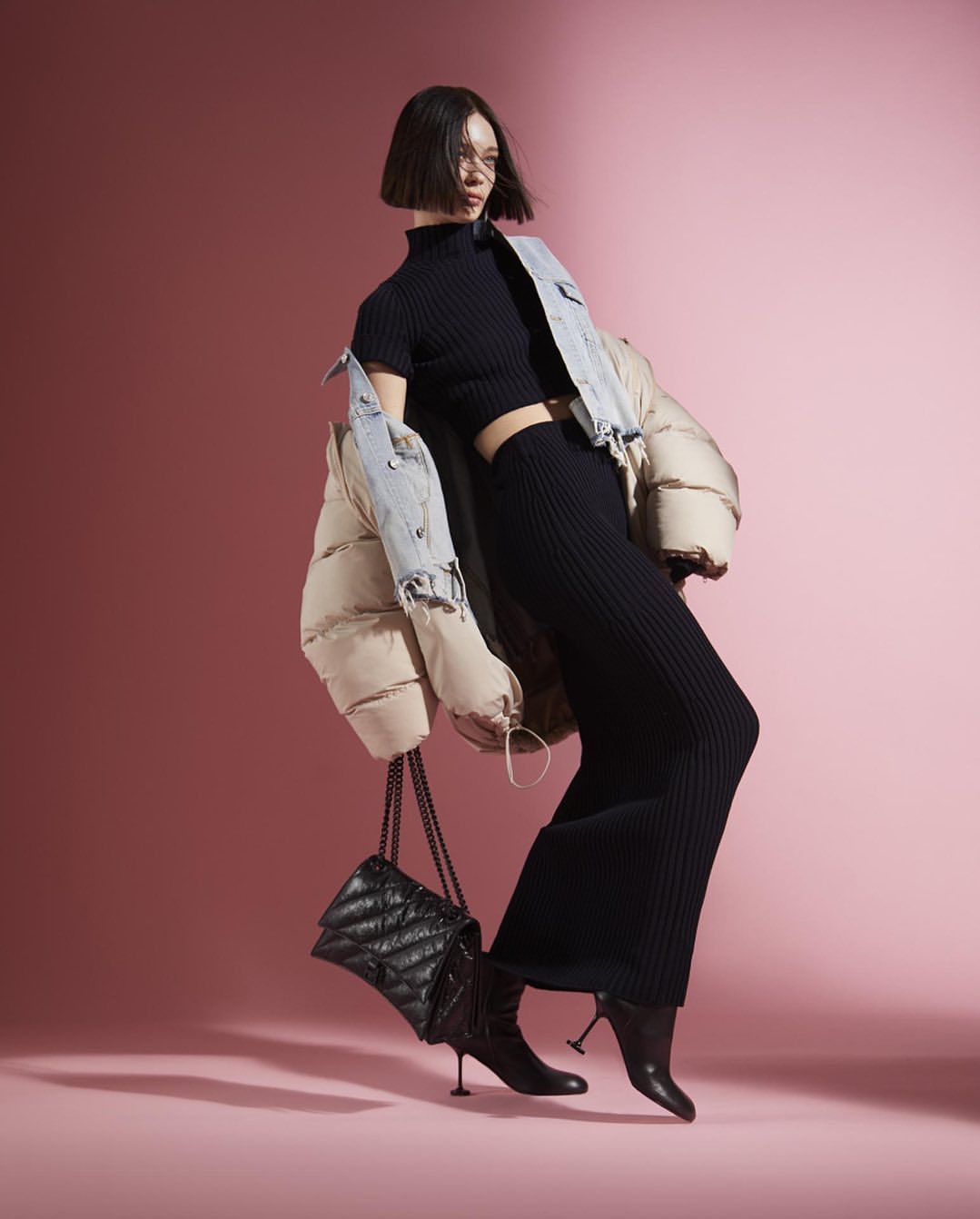 Fashionable woman in knitwear and a puffer jacket strikes a pose in a studio, showcasing a trendy black quilted handbag.