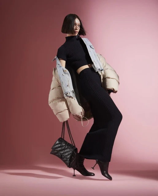 Fashionable woman in knitwear and a puffer jacket strikes a pose in a studio, showcasing a trendy black quilted handbag. 