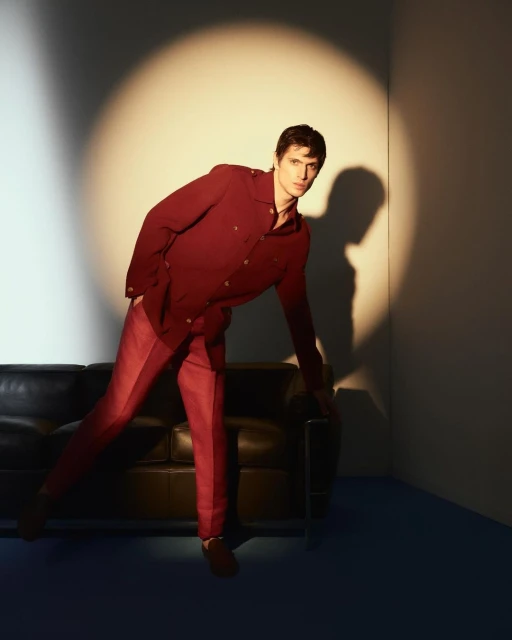 Stylish adult male poses indoors in a maroon suit jacket and pants, leaning on a modern black couch under studio spotlighting.
