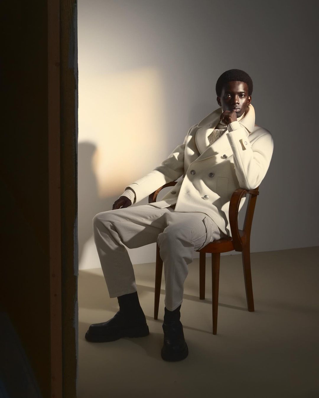 Elegant portrait of a Black man in a white double-breasted coat and light pants, sitting in a chair in front of a simple, light-colored wall.