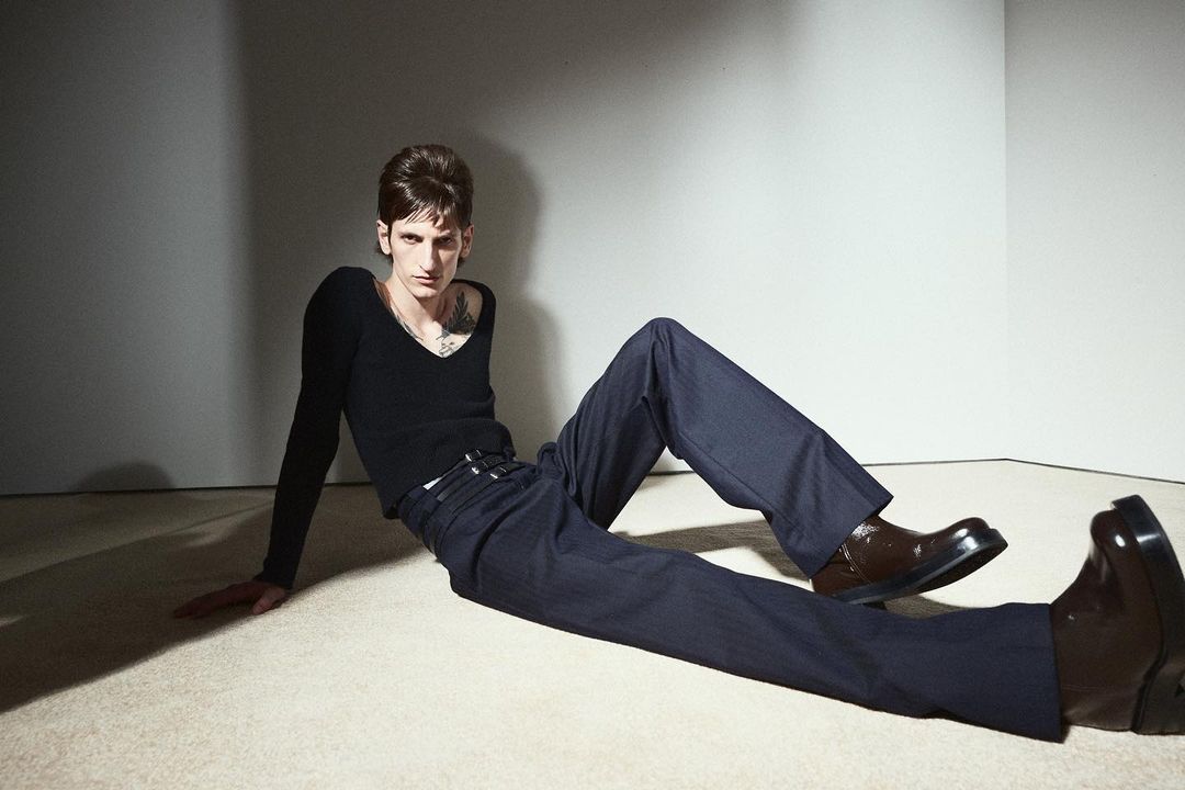 Adult male fashion model with dark hair poses on the floor in a long-sleeved black V-neck shirt, dark blue pants, and shiny brown shoes.