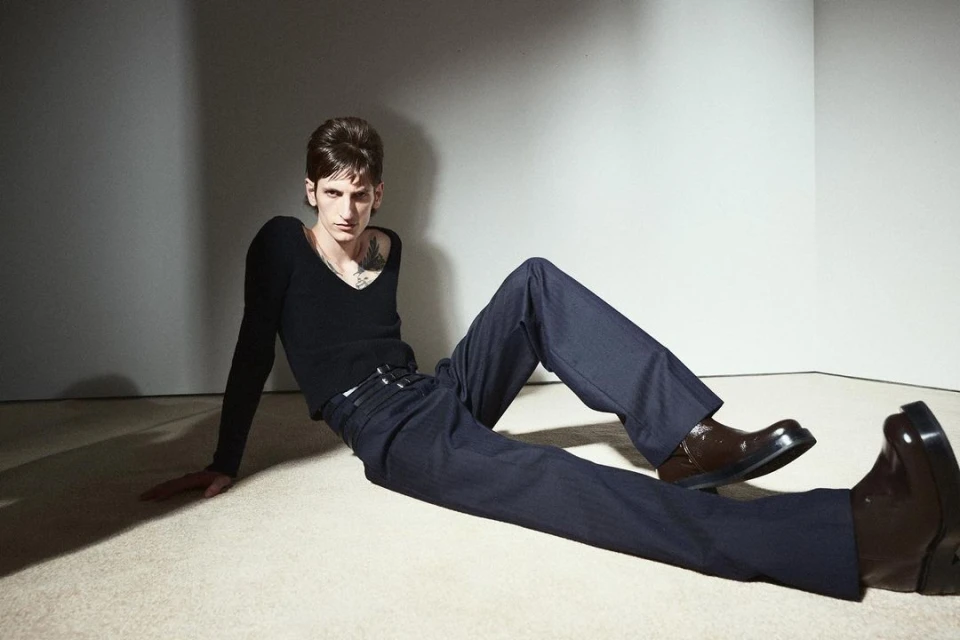 Adult male fashion model with dark hair poses on the floor in a long-sleeved black V-neck shirt, dark blue pants, and shiny brown shoes.