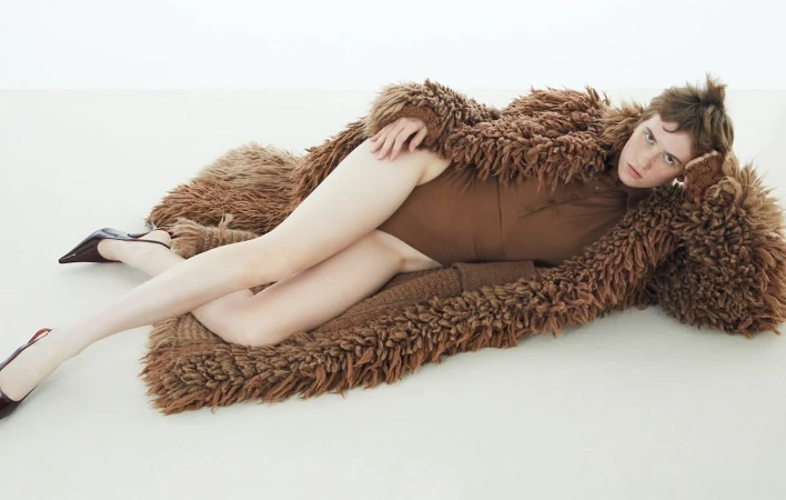 Fashion shot of a woman reclining on a brown, shaggy rug wearing a brown top and black heels in a brightly lit photo studio.