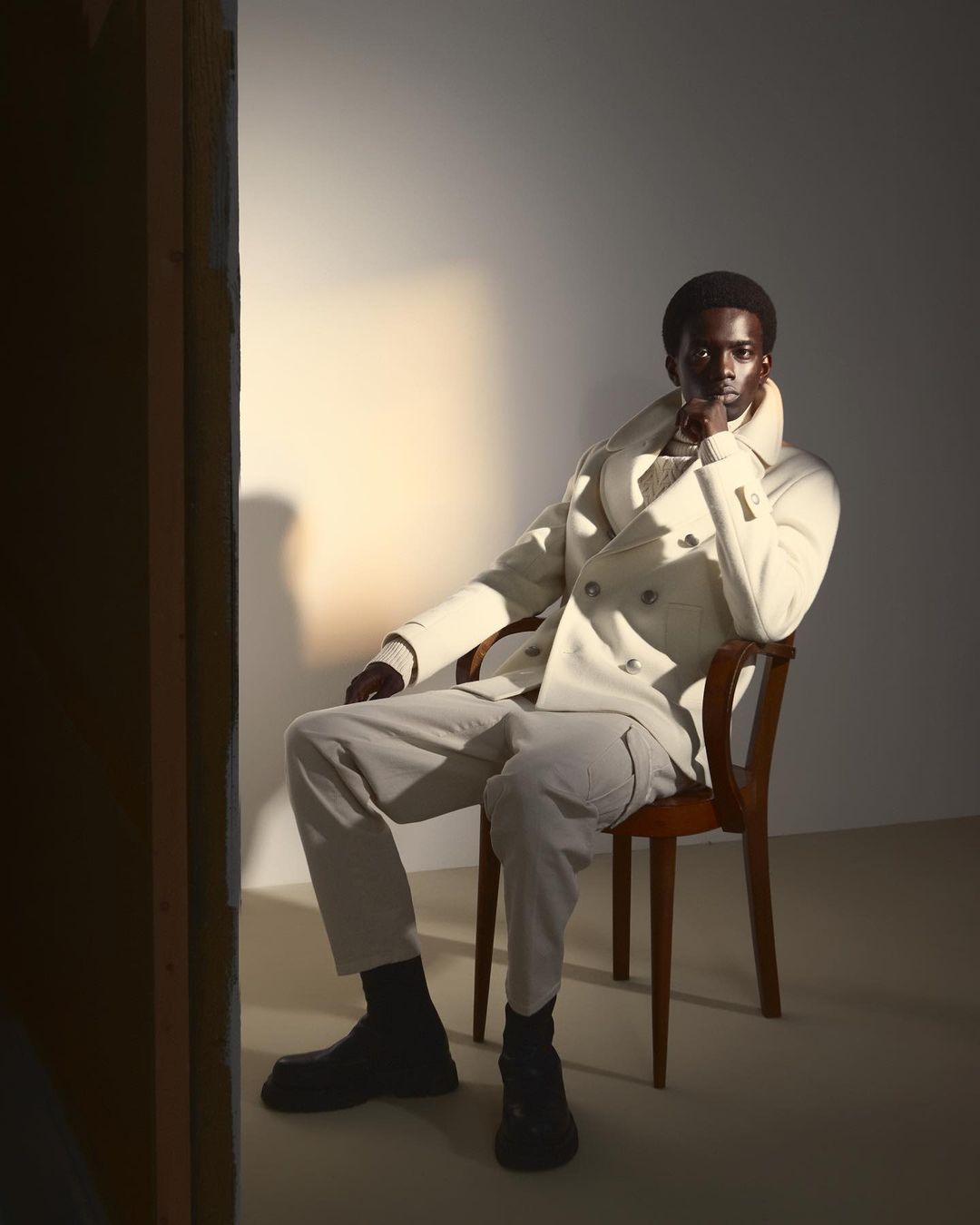 An adult male of African descent poses while seated in a studio, illuminated by a soft spotlight, wearing a cream double-breasted coat and light pants.