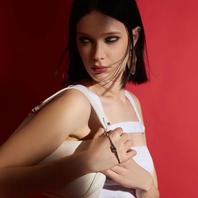 Brunette woman with smokey eye makeup wearing a white top and holding a purse in front of a red backdrop with side lighting.