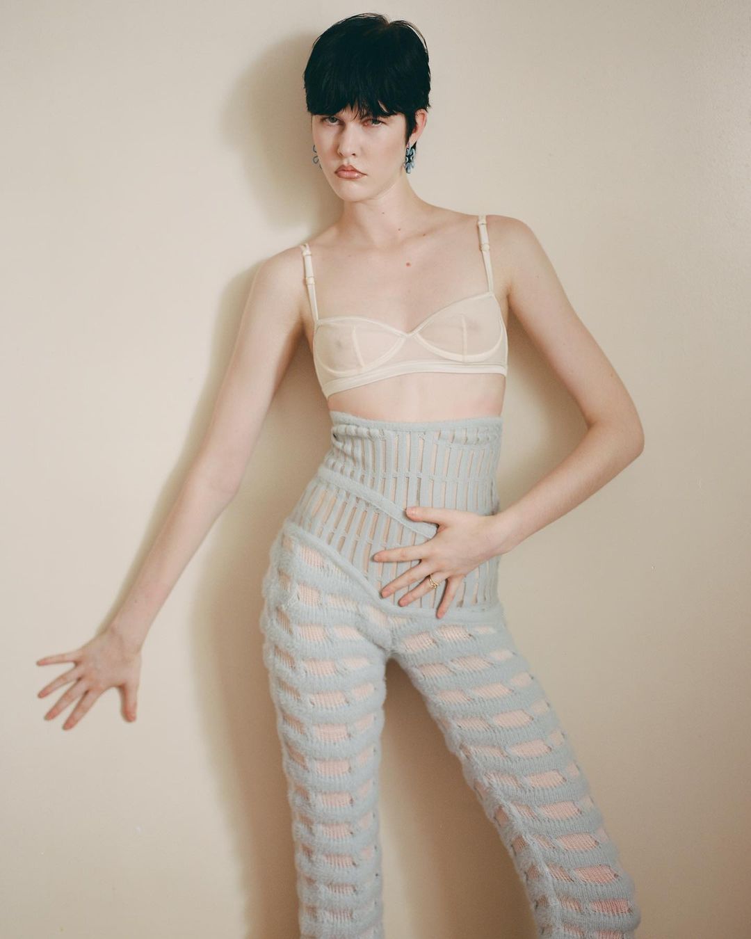 Fashionable woman with short dark hair poses in a beige bra and blue knit pants against a neutral wall in a studio shot.