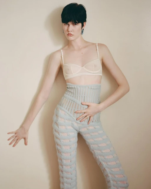 Fashionable woman with short dark hair poses in a beige bra and blue knit pants against a neutral wall in a studio shot.