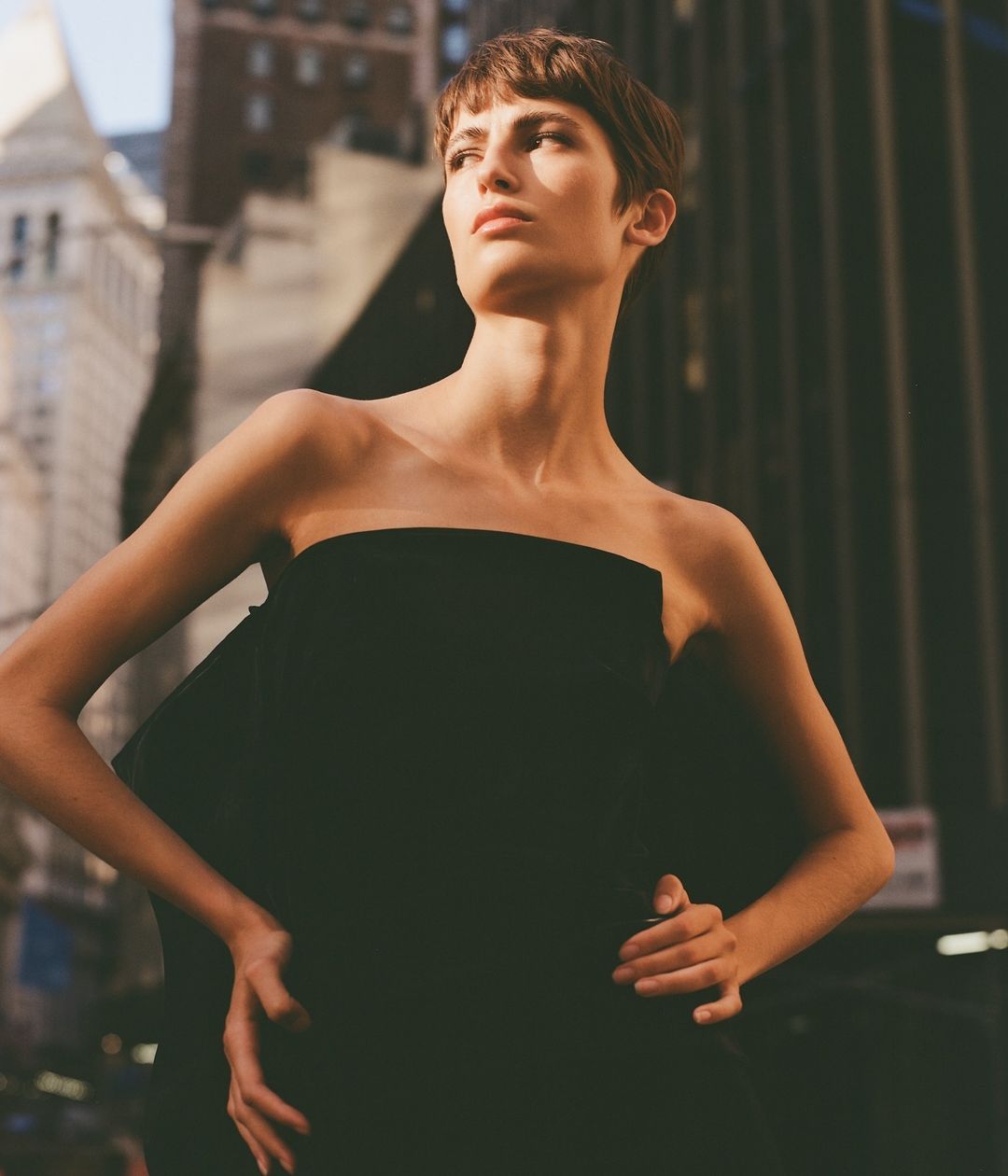 Model with short brown hair wearing a strapless black dress posing outdoors against an urban architectural street backdrop