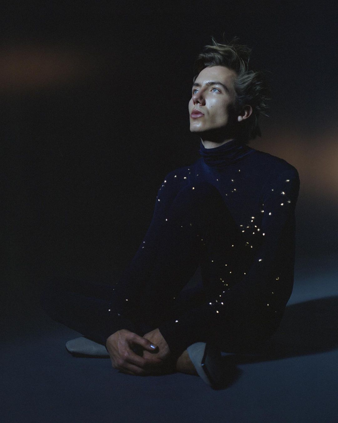 Portrait of a young adult male with light skin and spiky blonde hair, wearing a black turtleneck sweater, seated on a blue surface, looking up.