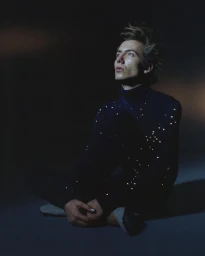 A man wearing a dark turtleneck with sparkling embellishments seated on the floor against a dark background