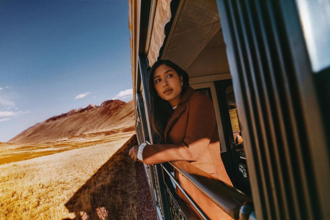 Woman in a brown coat looks out from a vintage train window, mountains and fields visible in the distance on a sunny day.