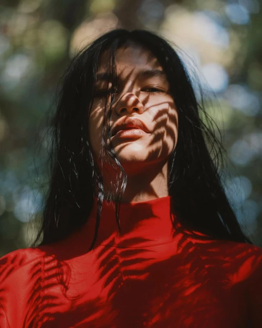 Close-up portrait of a young woman with dark hair, red top, and leaf shadows across her face, set against a blurred leafy background.